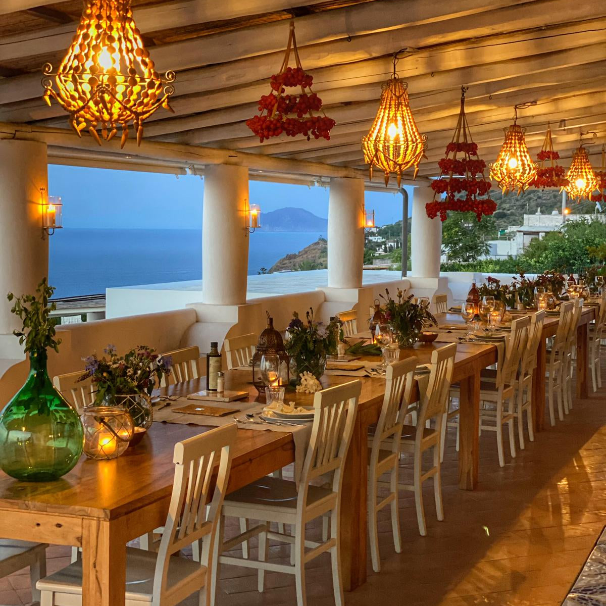An indoor dining area with wooden tables and chairs, featuring chandeliers with a predominantly red hue, and large windows in the background showcasing a scenic view.
